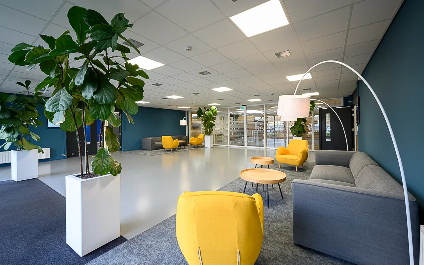 Moderne wachtruimte aan de Essebaan met grijze banken, gele stoelen, hoge planten en een strakke inrichting.