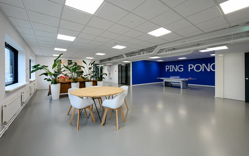 Moderne kantoorruimte met een ronde vergadertafel, grote planten en een pingpongtafel tegen een blauwe muur met de tekst "PING PONG".