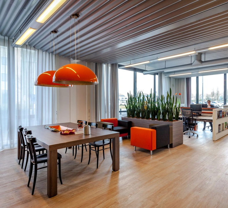 Modern kantoorinterieur met houten vloer, grote oranje hanglampen, een vergadertafel en een loungegedeelte met planten en zitbanken.