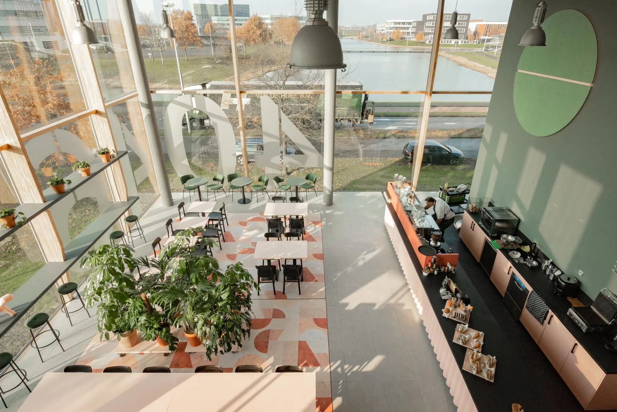 Modern ingerichte horecaruimte aan de Hondiuslaan met grote raampartijen, kleurrijke vloer, planten en een open koffiebar.