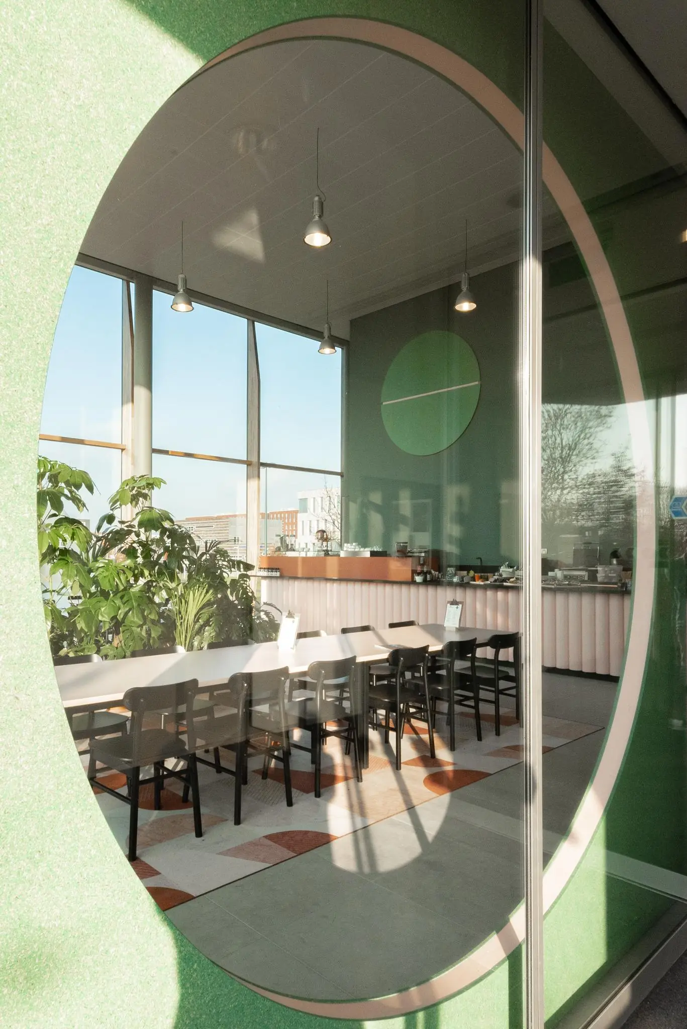Modern ingericht café aan de Hondiuslaan met lange tafel, zwarte stoelen, veel daglicht en groene accenten.