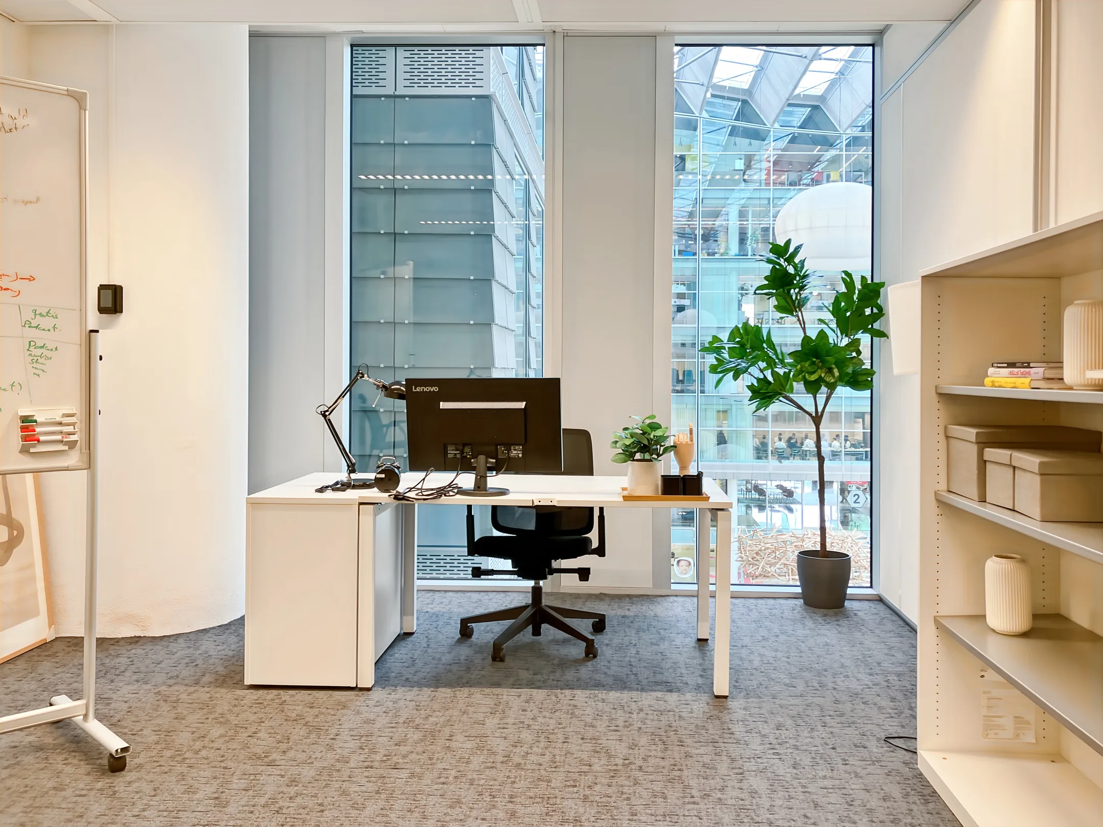 Een moderne kantoorruimte met een bureau, computer, planten en uitzicht op een glazen gebouw.