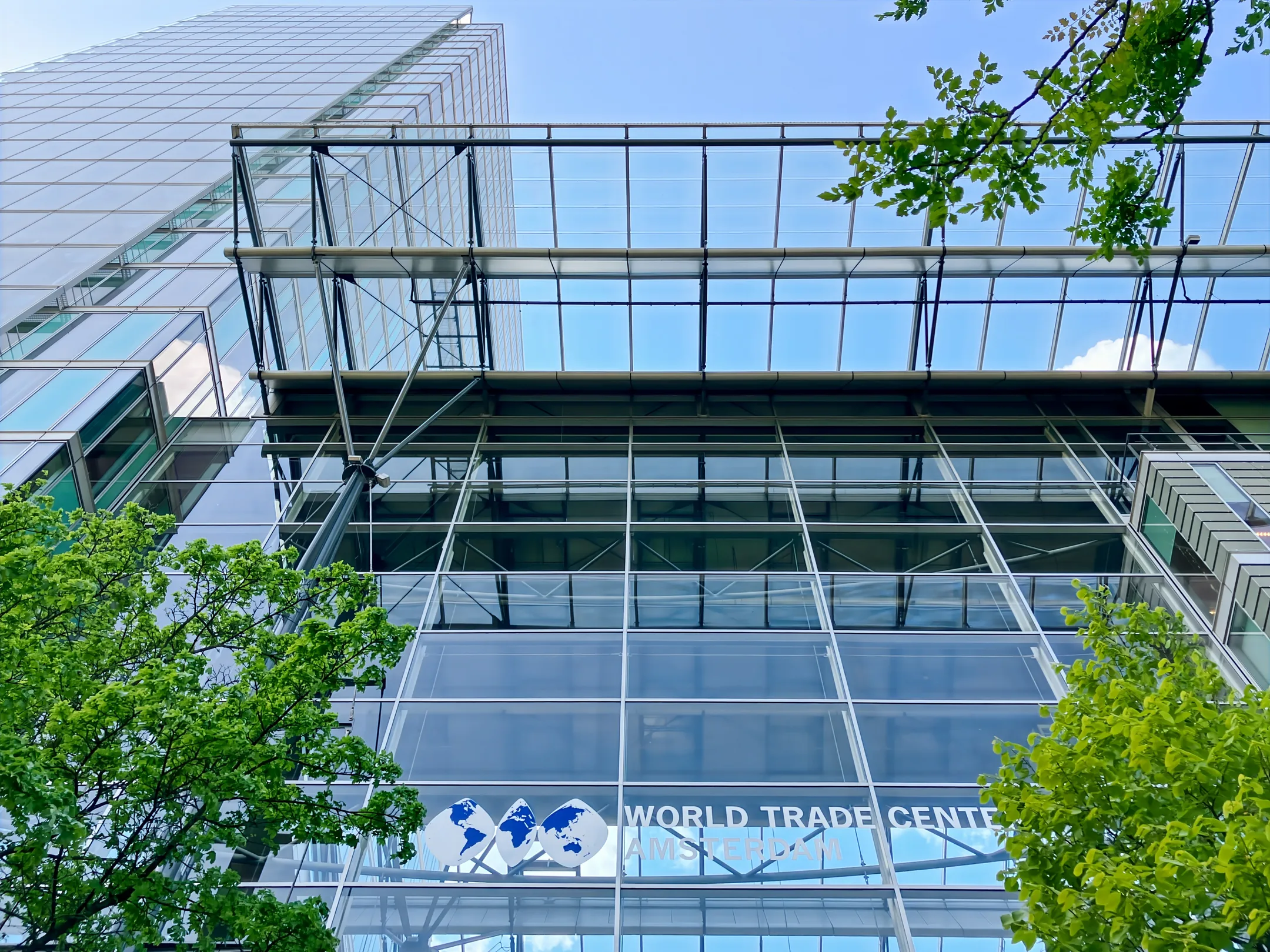 Het World Trade Center Amsterdam gebouw met glazen gevel, gezien van onderaf te midden van groene bomen.