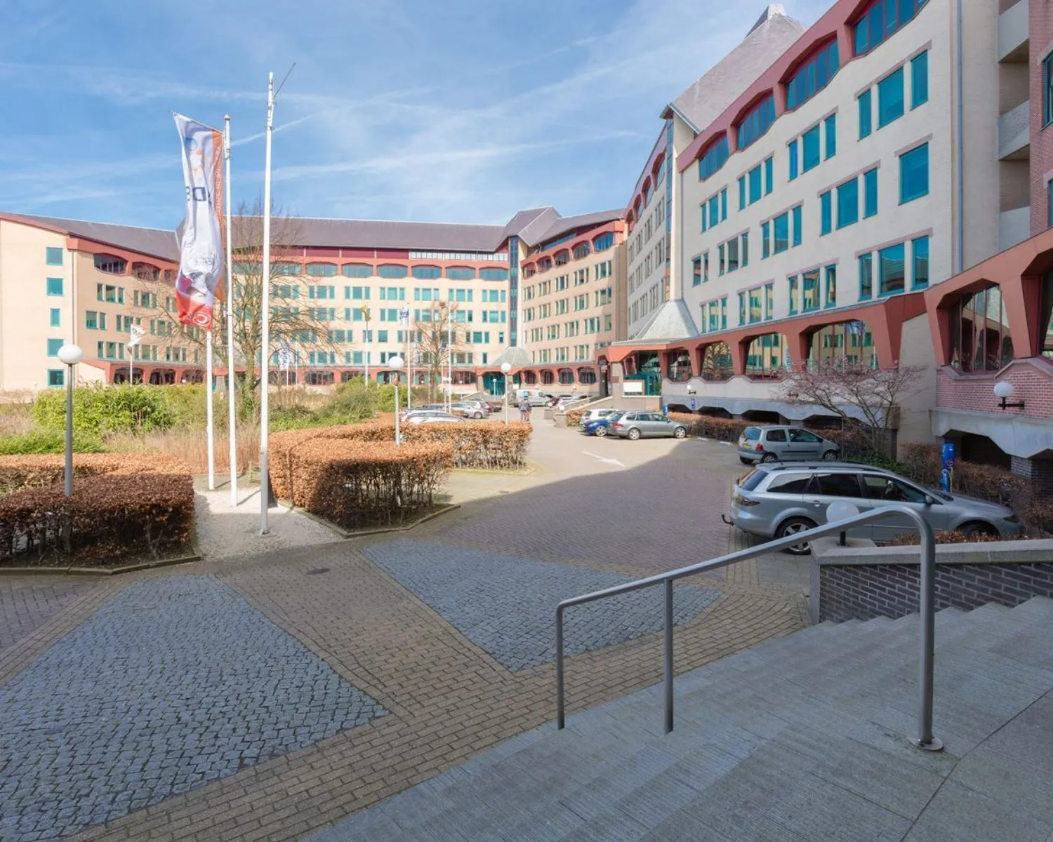 Vooraanzicht van een groot kantoorgebouw met parkeerplaatsen en vlaggenmast op een zonnige dag.