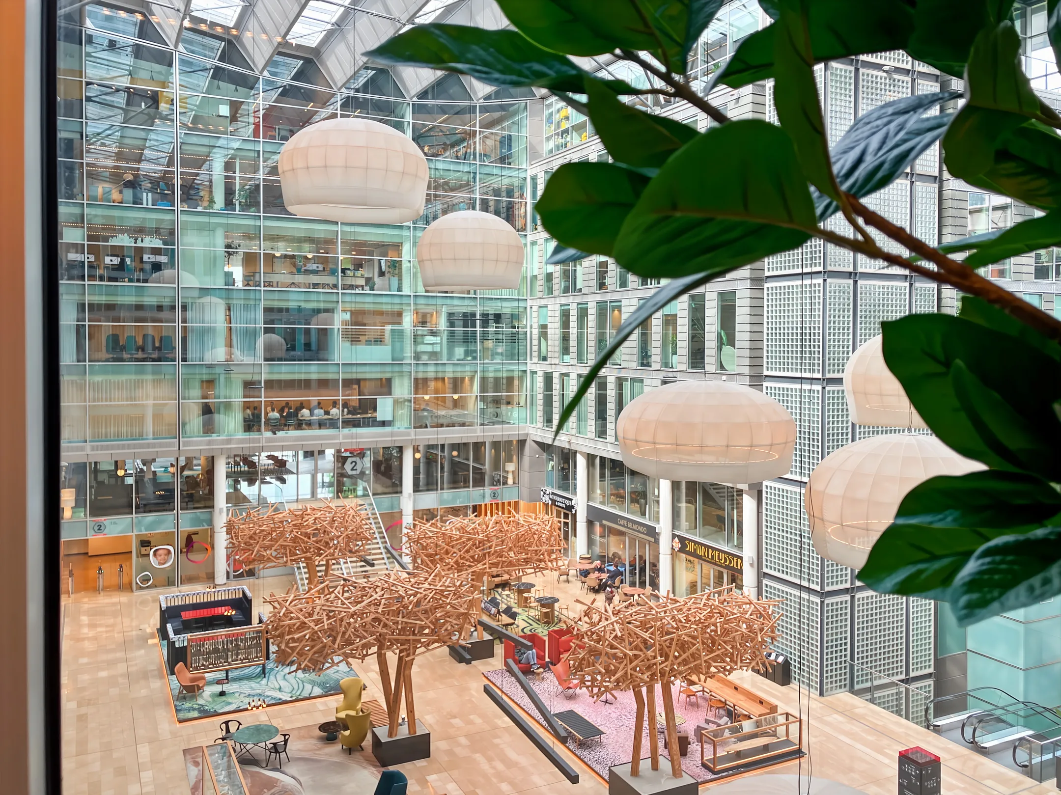 Modern atrium met glazen wanden, grote hanglampen en zitgedeeltes met decoratieve houten structuren.