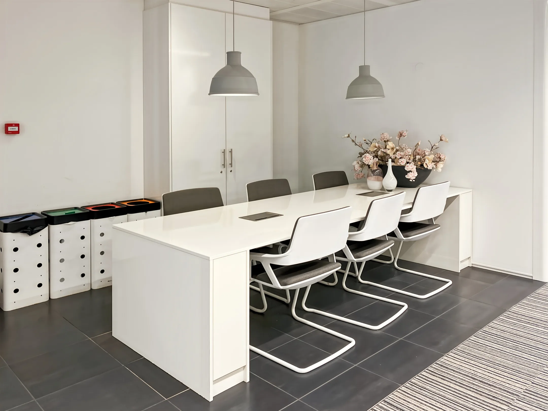 Een moderne vergaderruimte met een witte tafel, zes stoelen, hanglampen en en een grote vaas met bloemen.