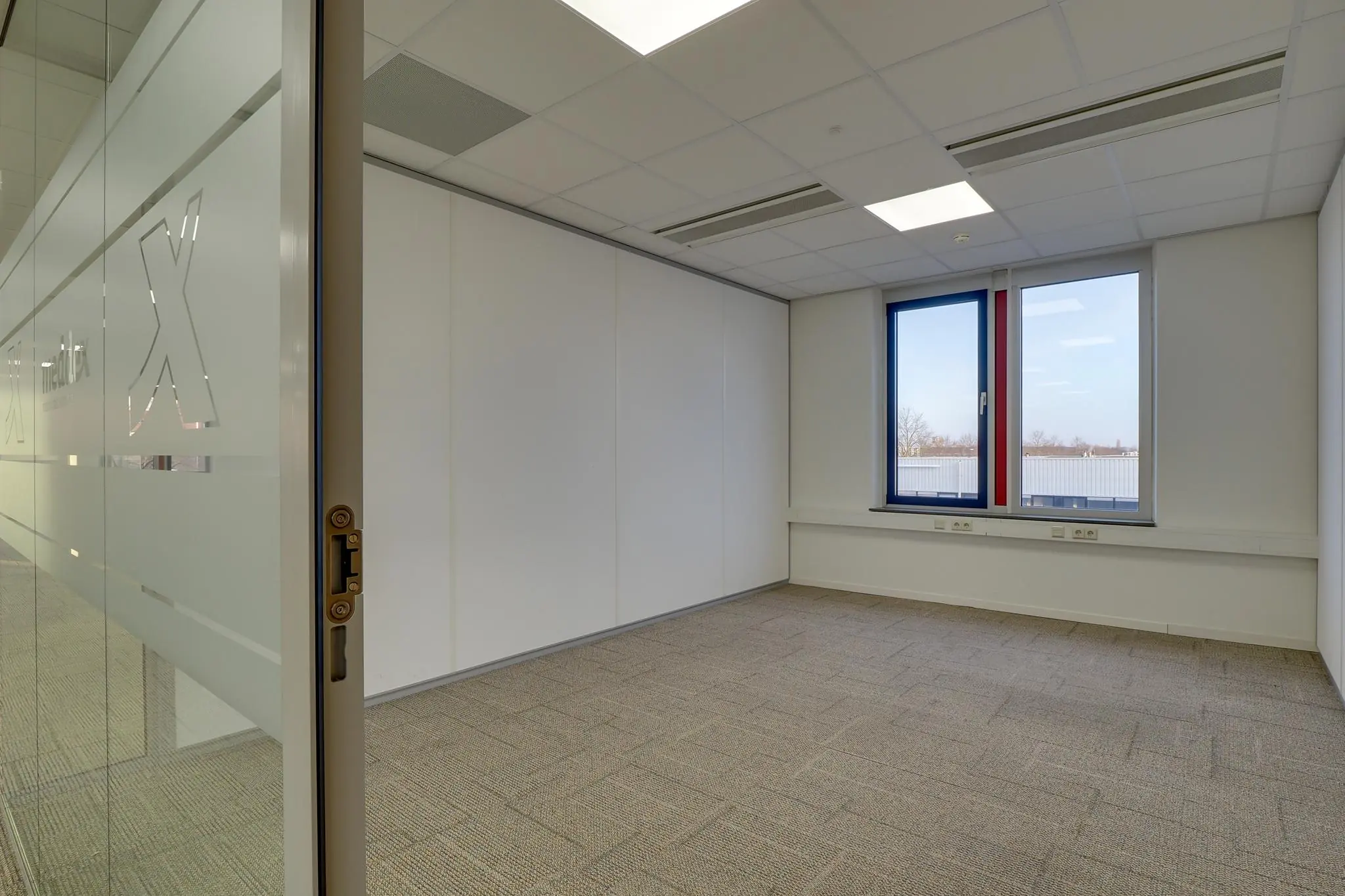 Leeg kantoor met glazen deur, wit plafond, tapijtvloer en een raam met uitzicht op een bedrijventerrein aan de Rijnzathe.