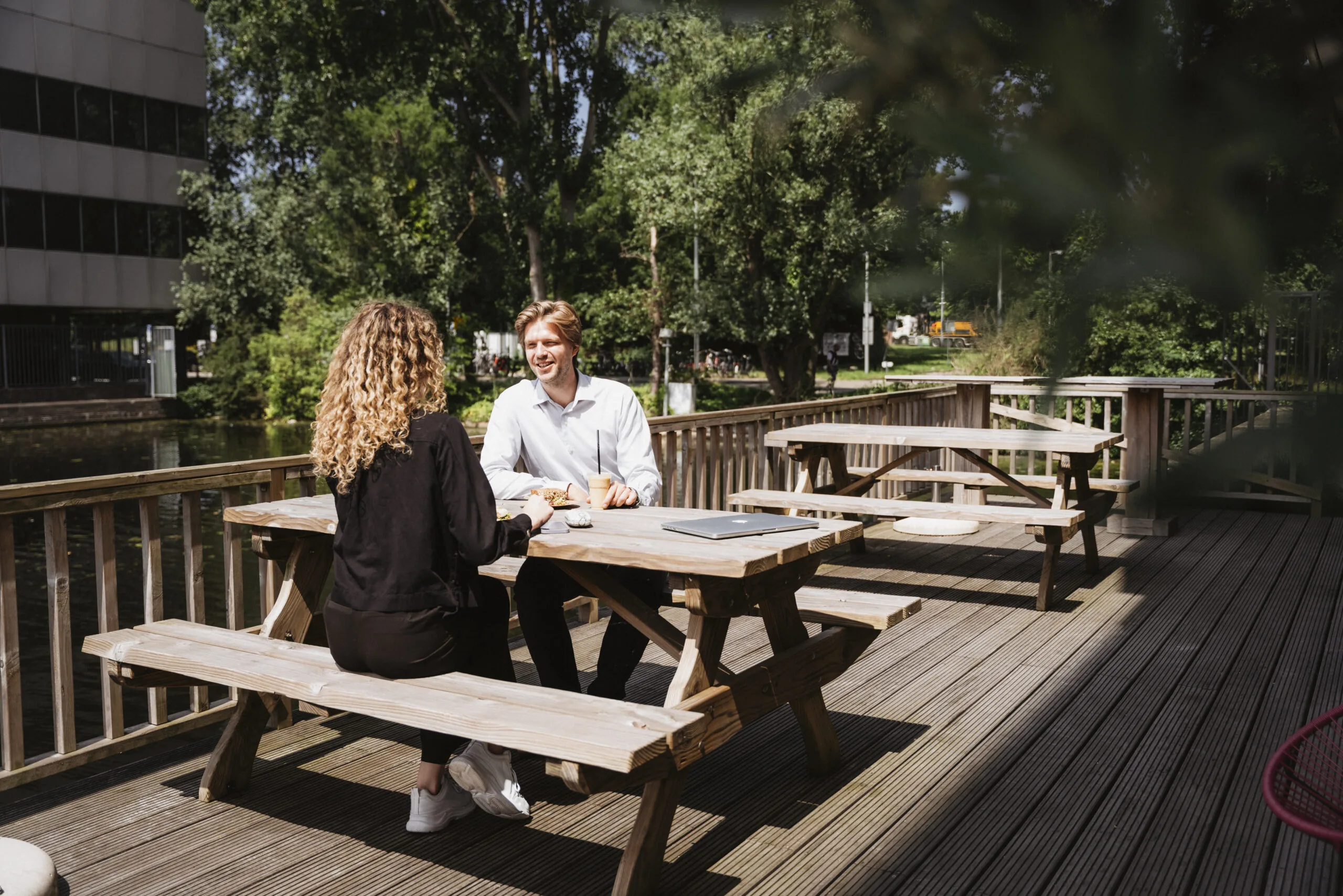 Twee mensen zitten op het terras van HNK Amsterdam Schinkel 15 aan een picknicktafel te lunchen en praten in de zon.