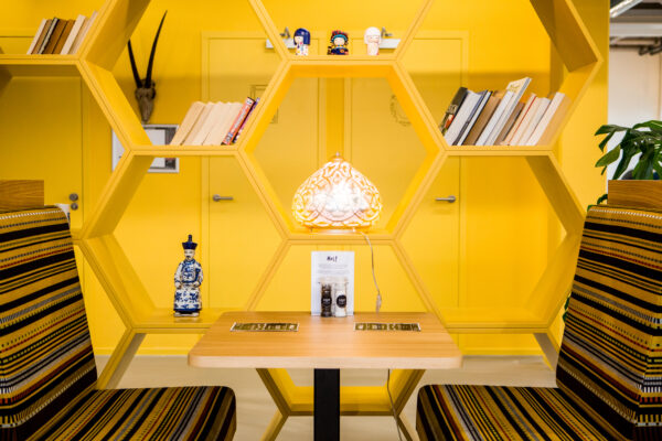 Geel ingerichte zithoek in HNK Amsterdam Zuidoost met bijenkorfvormige boekenplanken, decoratieve items en een verlichte tafellamp.