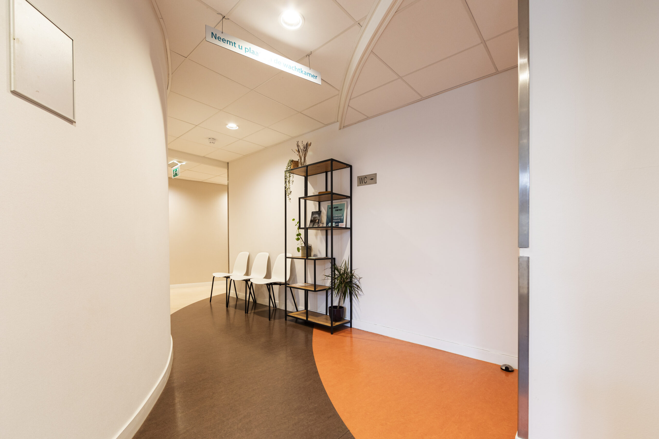 Wachtruimte met witte stoelen, een open kast met decoraties, en een oranje vloer.