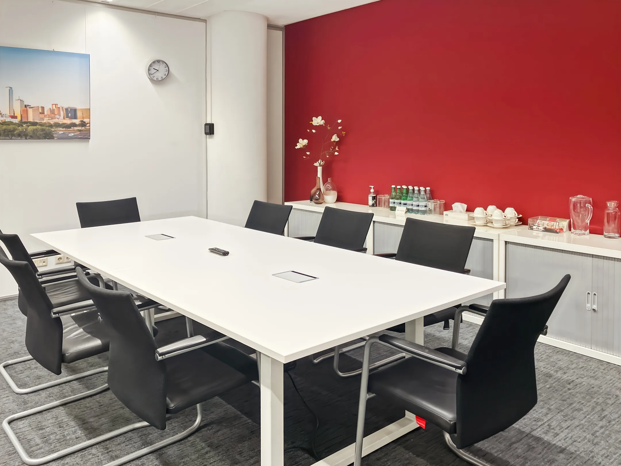 Een moderne vergaderruimte met een witte tafel, zes zwarte stoelen, en een rode muur met een kast vol drankjes en servies.