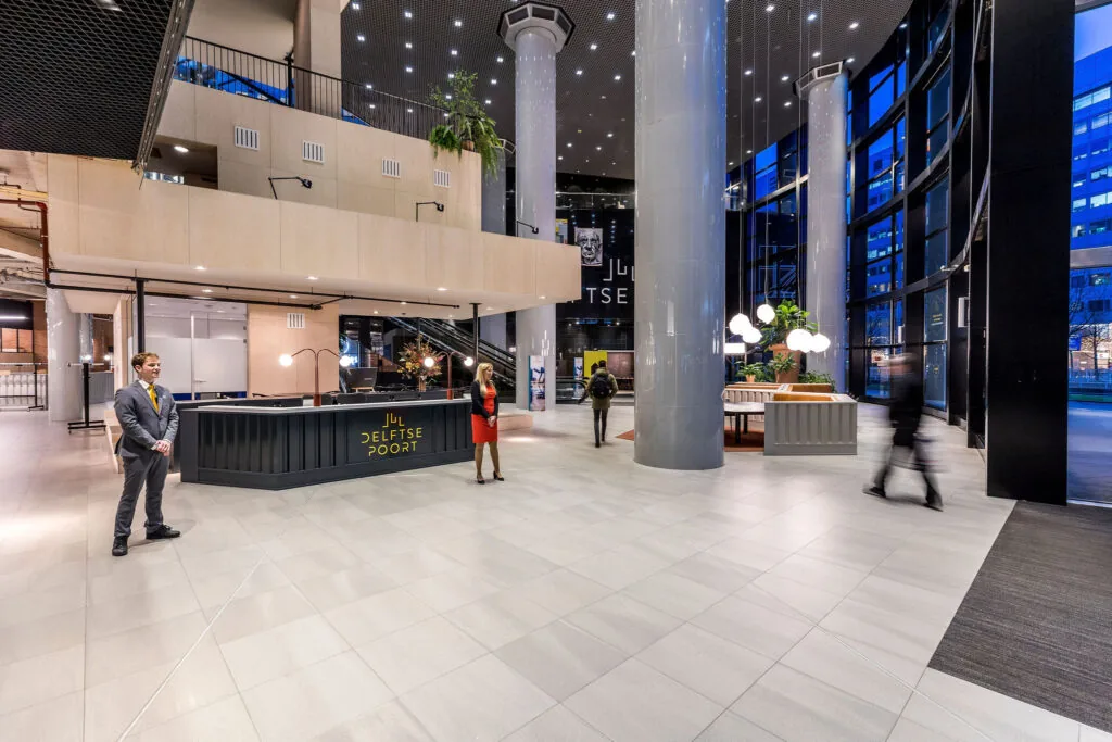 Moderne ontvangsthal van gebouw De Delftse Poort met balie, planten, verlichting en passanten.