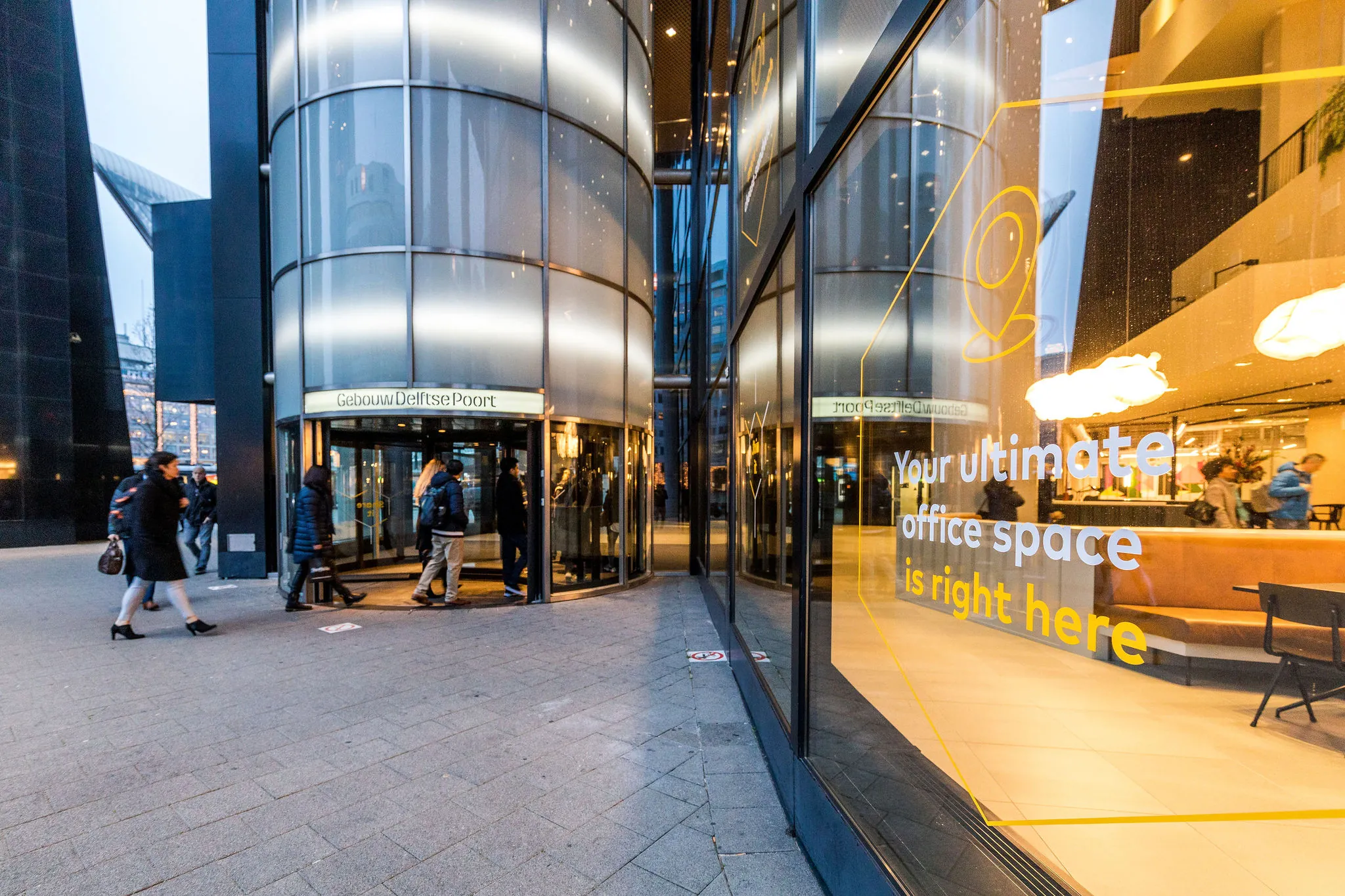 Ingang van het kantoorgebouw Delftse Poort in Rotterdam met mensen die naar binnen lopen en een etalageraam met promotietekst voor kantoorruimte.