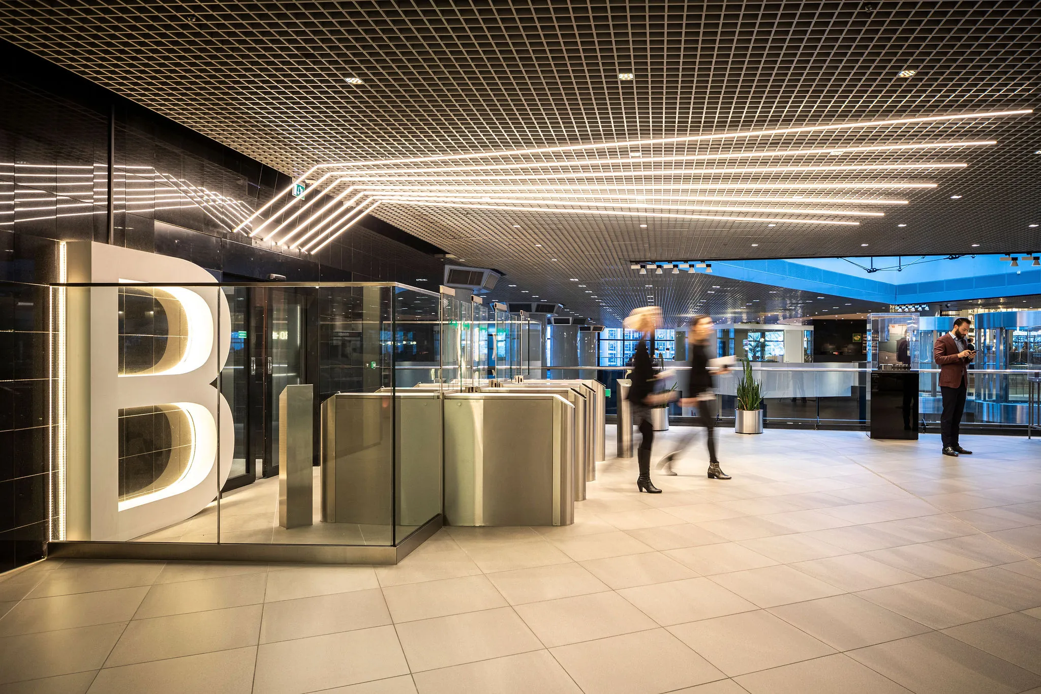 Moderne entreehal van het kantoorgebouw Weena met glazen tourniquets, een grote verlichte letter B en passerende mensen.