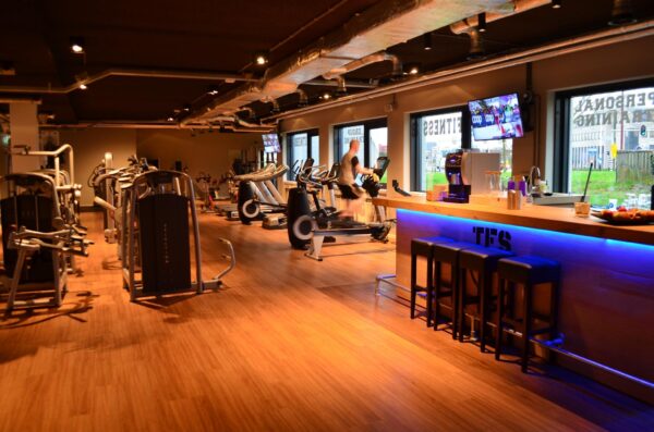 Moderne sportschool Nevelgaarde met fitnessapparatuur, een sporter op een crosstrainer en een sfeervolle bar met blauwe LED-verlichting.