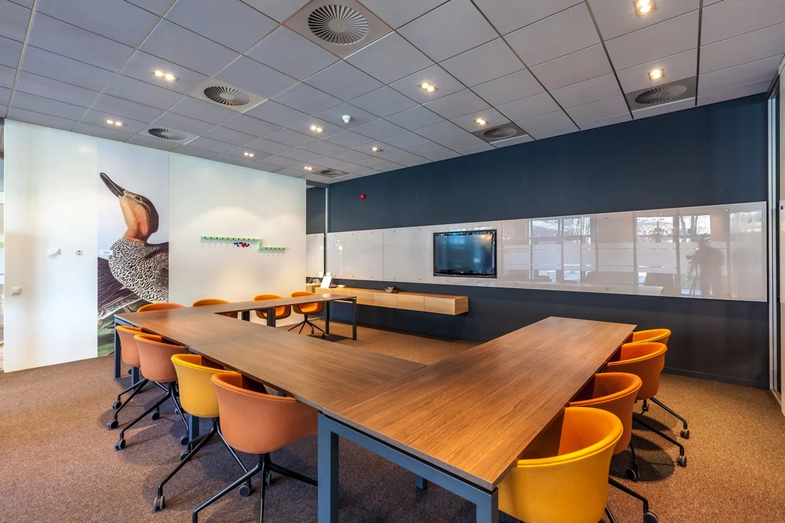 Vergaderruimte aan de Orteliuslaan met U-vormige tafel, oranje stoelen, flatscreen en wanddecoratie van een vogel.