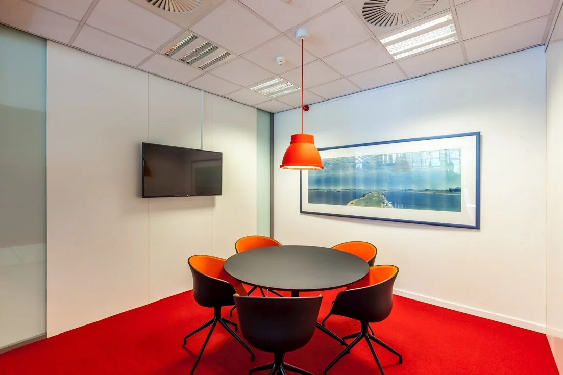 Vergaderruimte met een ronde zwarte tafel, vijf zwart-oranje stoelen, een rode hanglamp, flatscreen aan de muur en een panoramafoto van Orteliuslaan.