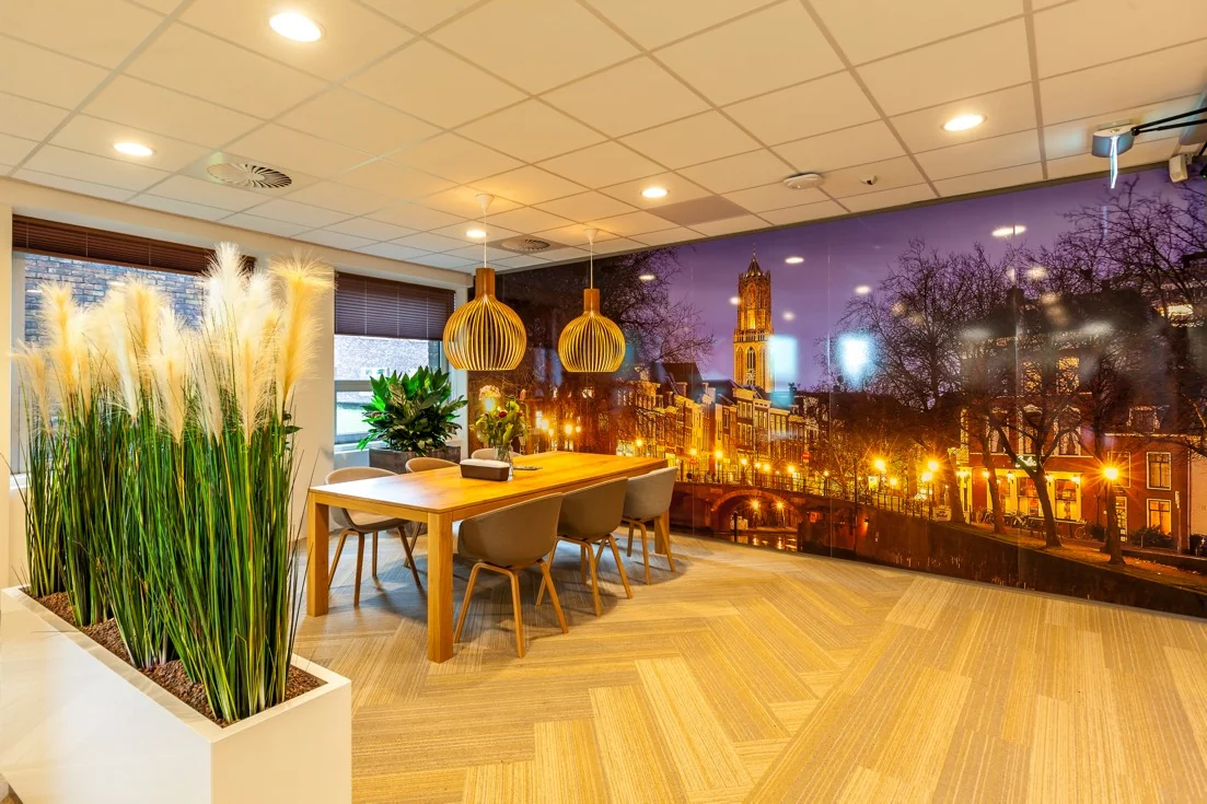 Moderne vergaderruimte met houten tafel, planten en een grote wandfoto van de Domtoren in Utrecht bij avond.