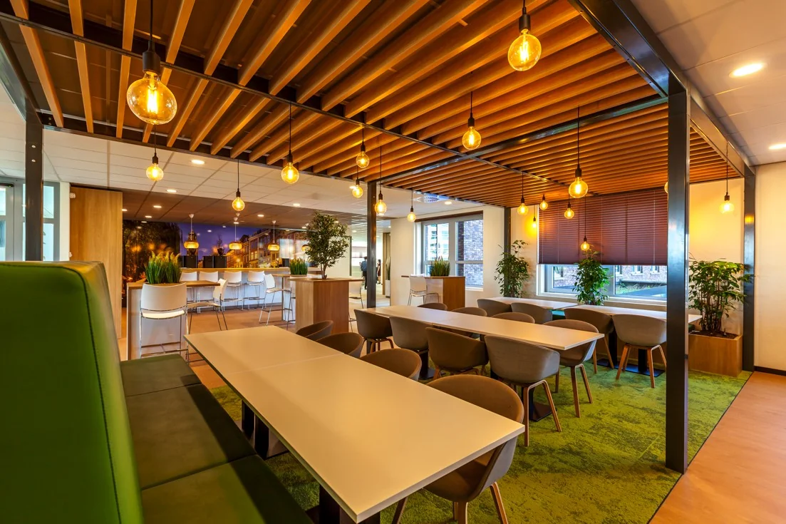 Moderne en sfeervolle kantoorkantine aan de Orteliuslaan met houten plafondaccenten, groene vloerbedekking en hanglampen.