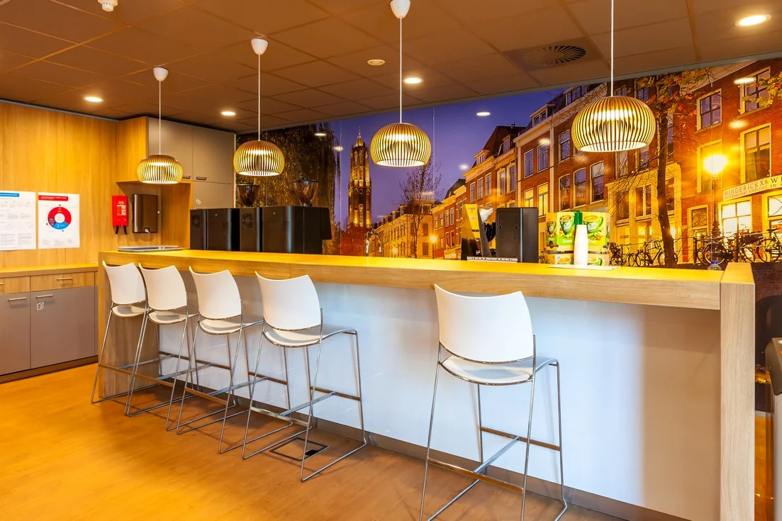 Moderne kantoor-koffiehoek met hoge stoelen, koffiemachines en een muurprint van de Orteliuslaan in Utrecht.