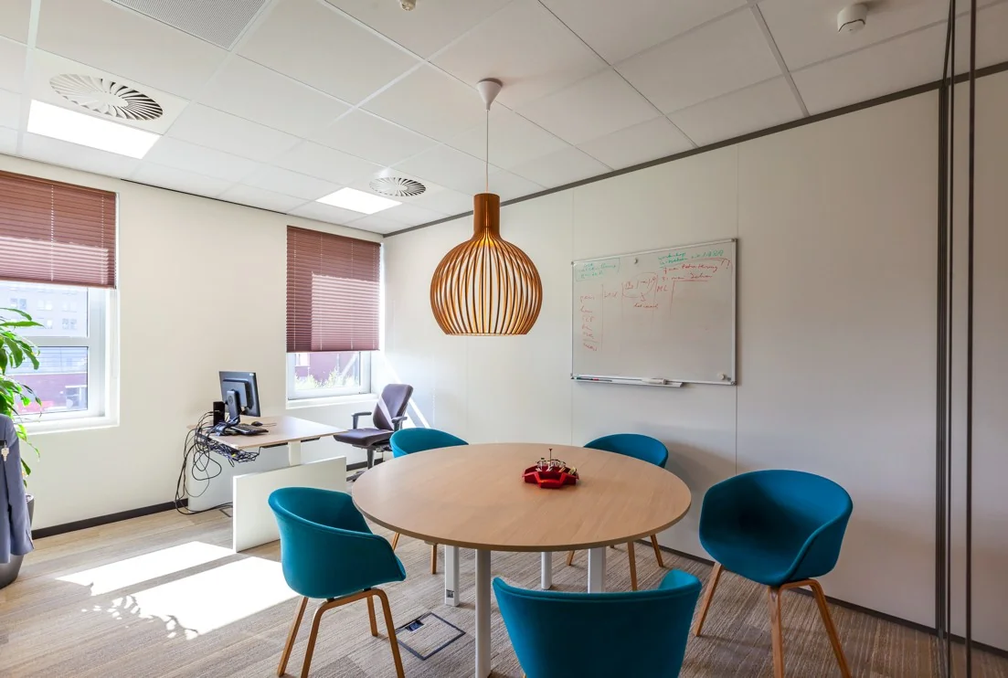 Moderne vergaderruimte aan de Orteliuslaan met ronde tafel, blauwe stoelen, whiteboard en werkplek bij het raam.