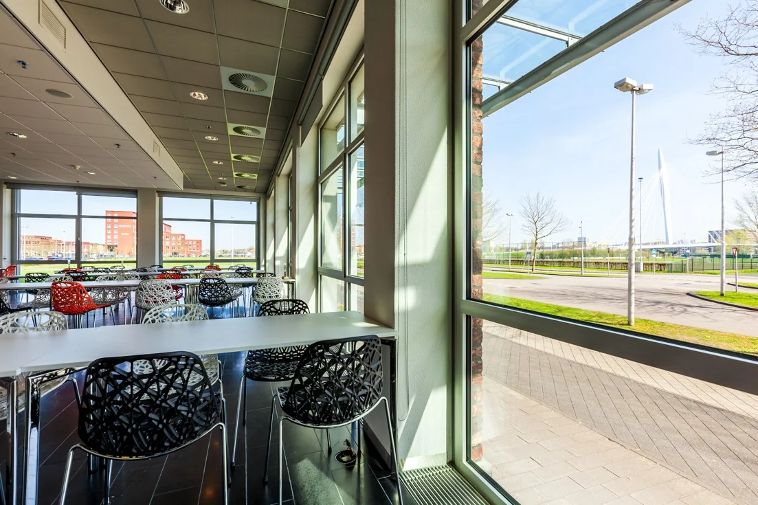 Moderne kantineruimte aan de Orteliuslaan met kleurrijke stoelen en uitzicht op een brug en kantoorpanden.