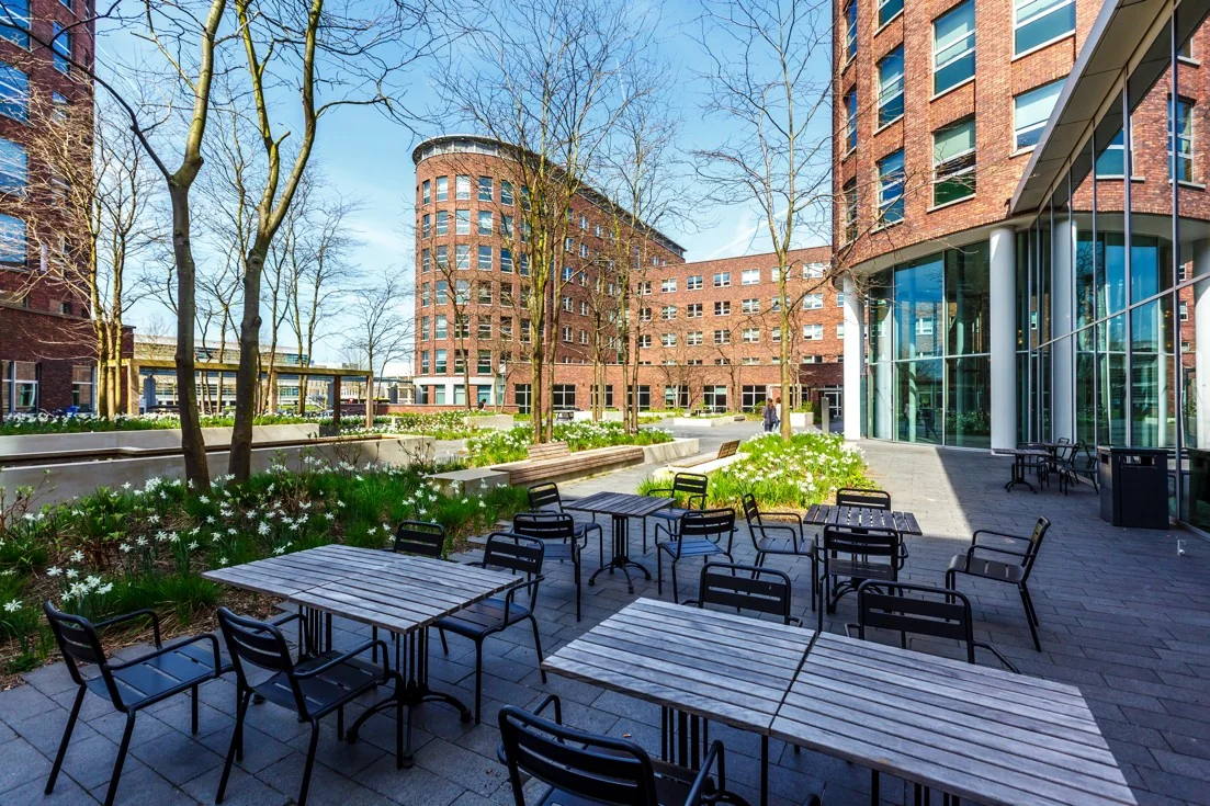 Buitenruimte met zitjes en bloemperken tussen moderne rode bakstenen kantoorgebouwen aan de Orteliuslaan.