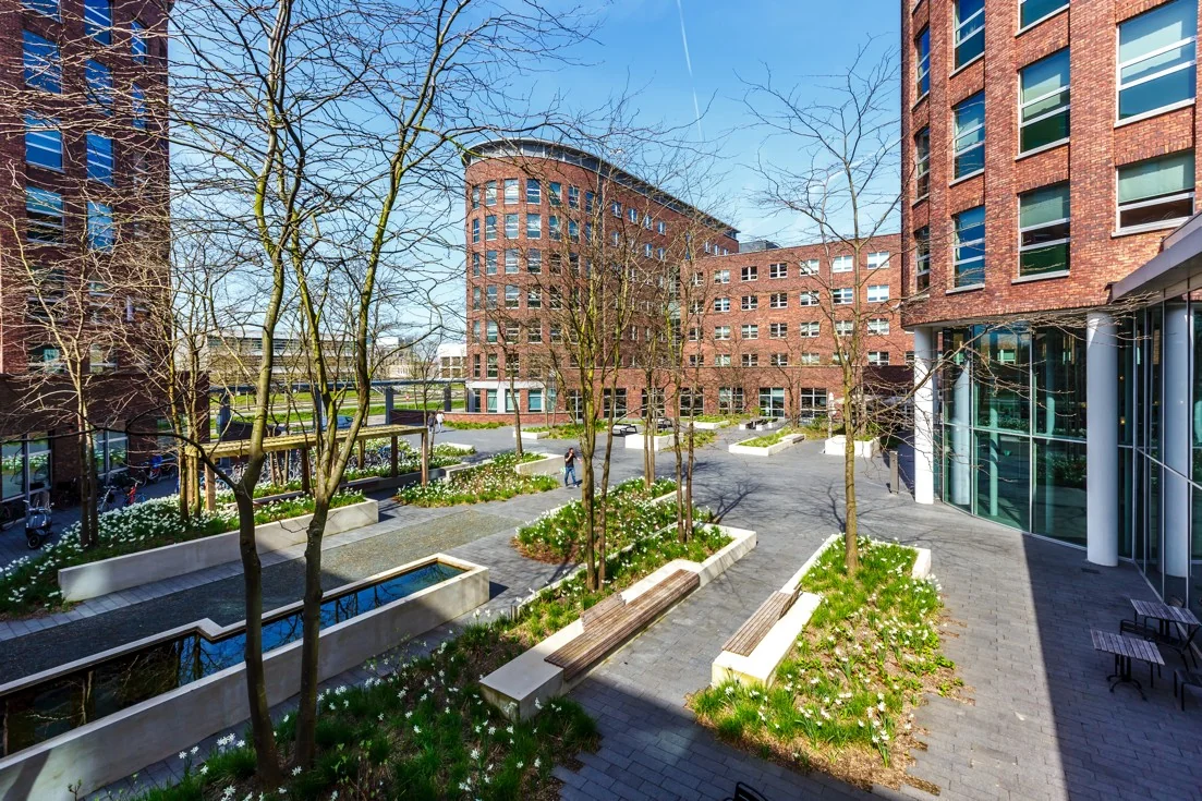 Binnenplein van kantoorpand aan de Orteliuslaan met rode bakstenen gebouwen, bomen, bankjes en bloemperken.
