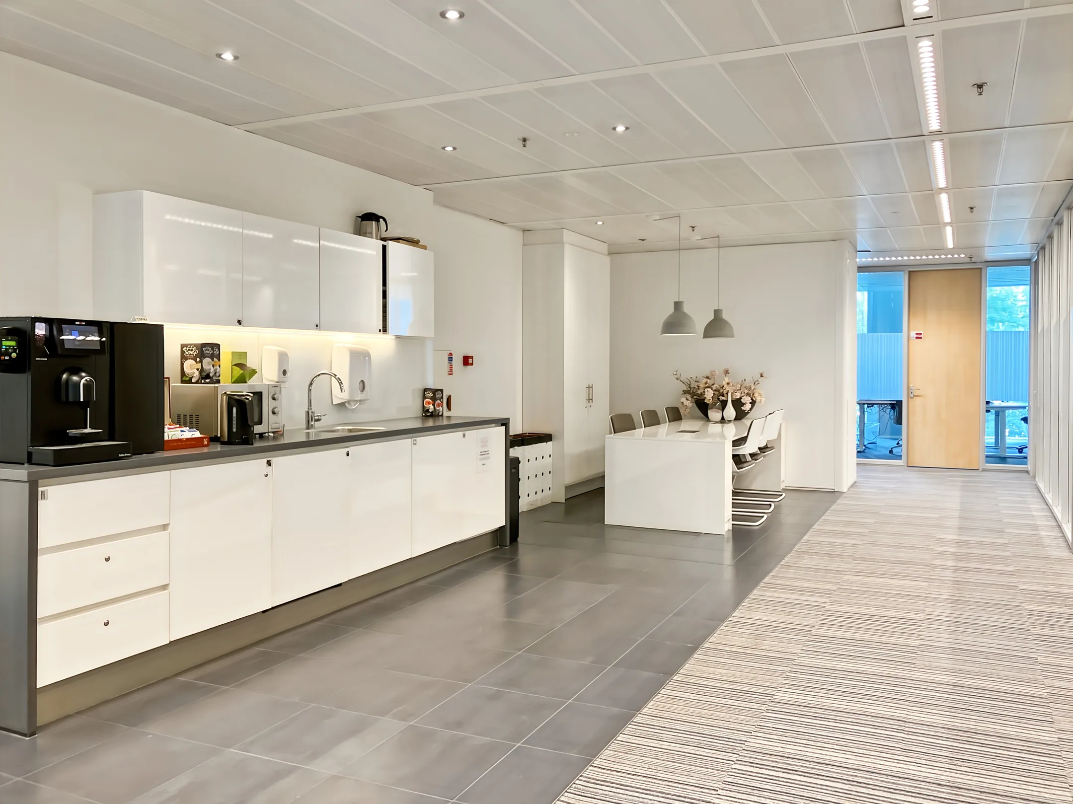 Een moderne kantoor keuken met een koffiezetapparaat, kasten en een eettafel met stoelen.