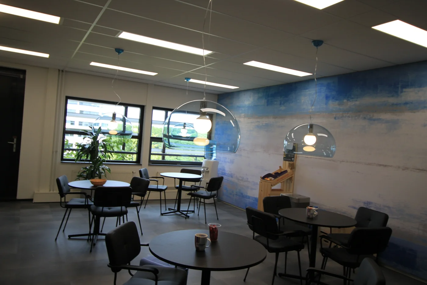 Moderne bedrijfskantine met ronde tafels, hanglampen en een blauw-wit geschilderde muur aan de Marconibaan.