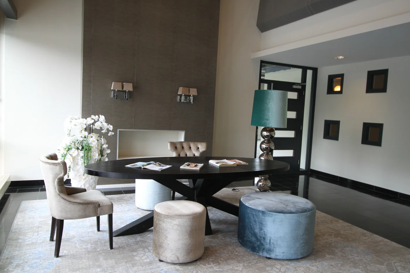Moderne wachtruimte met ronde zwarte tafel, diverse zitplaatsen, een grote vloerlamp en decoratieve bloemen op een kleed.