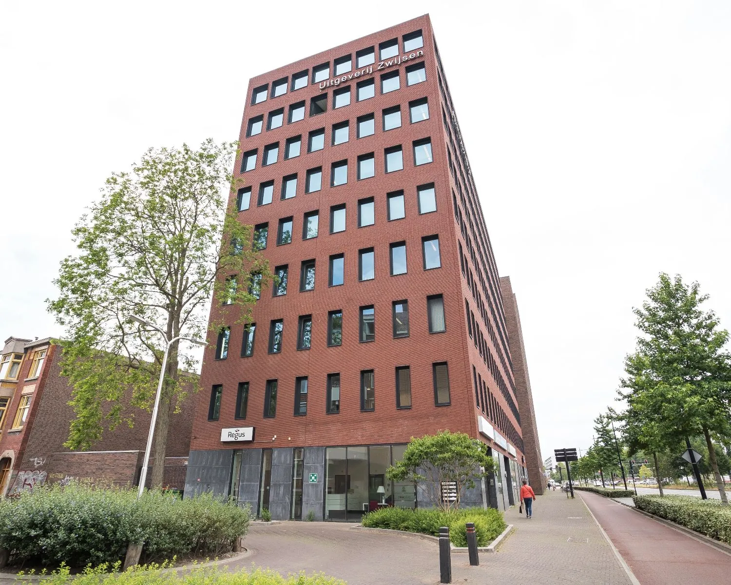 Hoog rood bakstenen kantoorgebouw van Uitgeverij Zwijsen aan een straat met bomen en fietspad.