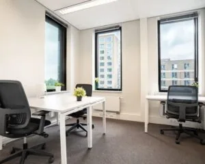Licht en modern ingericht kantoor met vier werkplekken, grote ramen en uitzicht op nabije flatgebouwen.