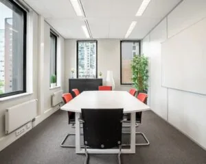 Vergaderruimte met een witte tafel, zes stoelen en grote ramen met uitzicht op omliggende gebouwen.