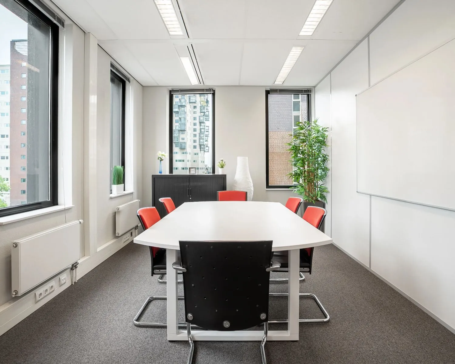 Vergaderruimte met een witte tafel, zes stoelen en grote ramen met uitzicht op omliggende gebouwen.