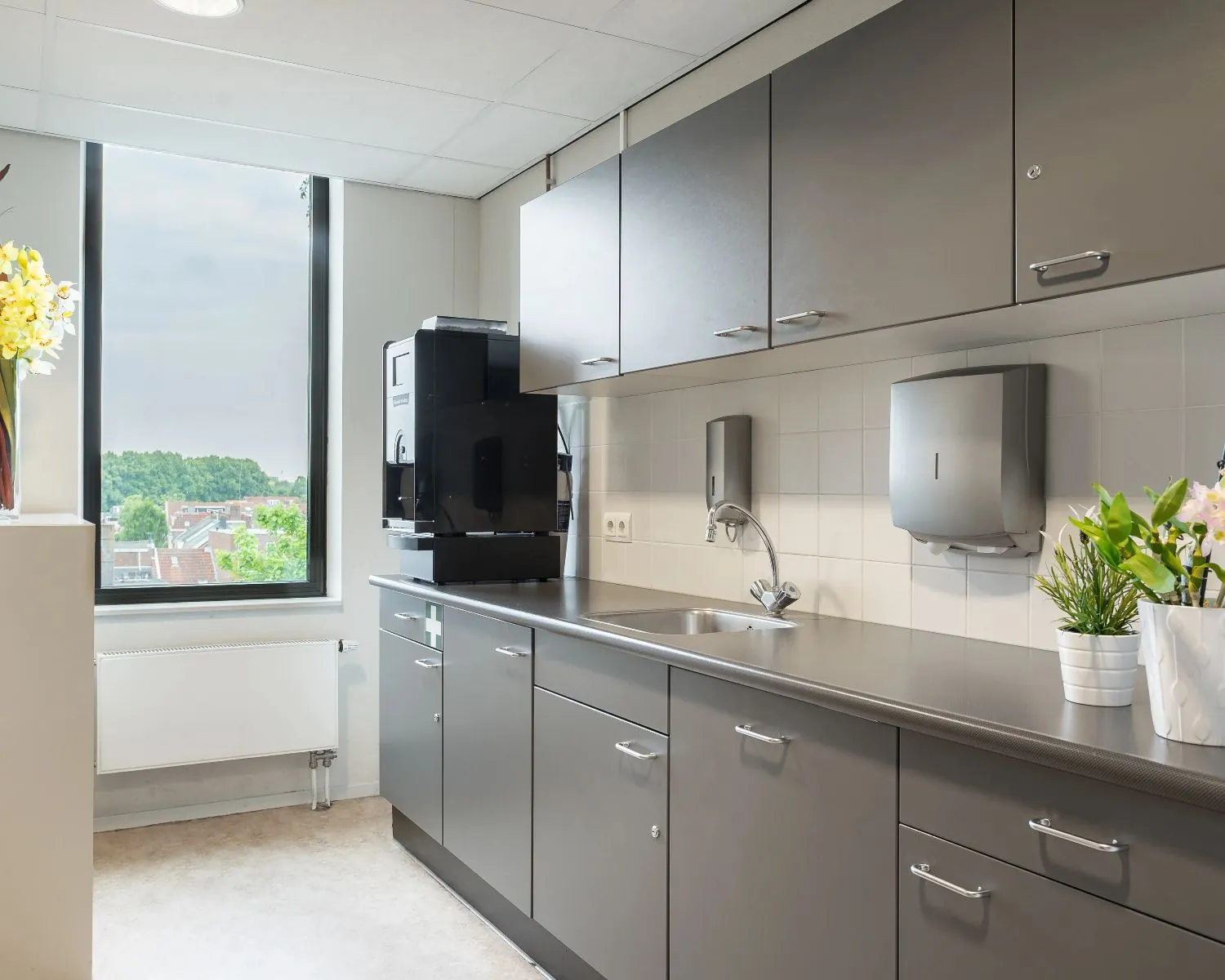 Moderne pantry met grijze keukenkastjes, een koffieautomaat en uitzicht op een stad met groen.