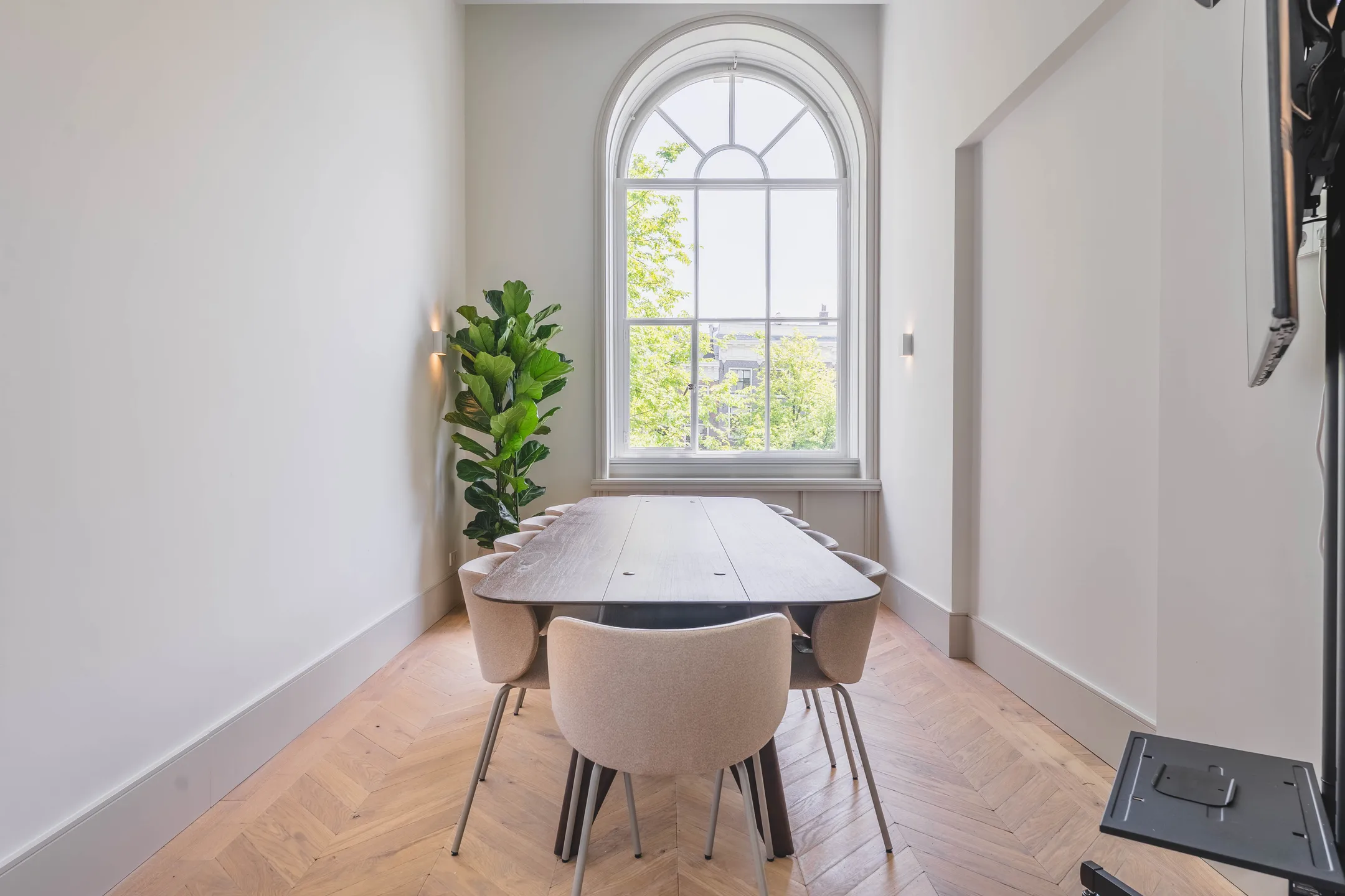 Een lichte vergaderruimte met een lange tafel, gestoffeerde stoelen, een grote boogramen en een groene plant in de hoek.