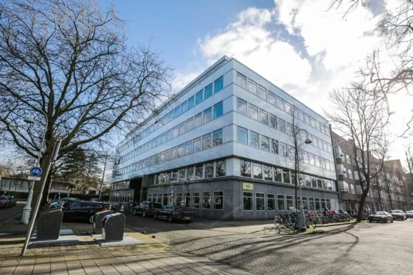 Modern kantoorgebouw op een straathoek met geparkeerde auto's en fietsen, onder een heldere blauwe lucht.