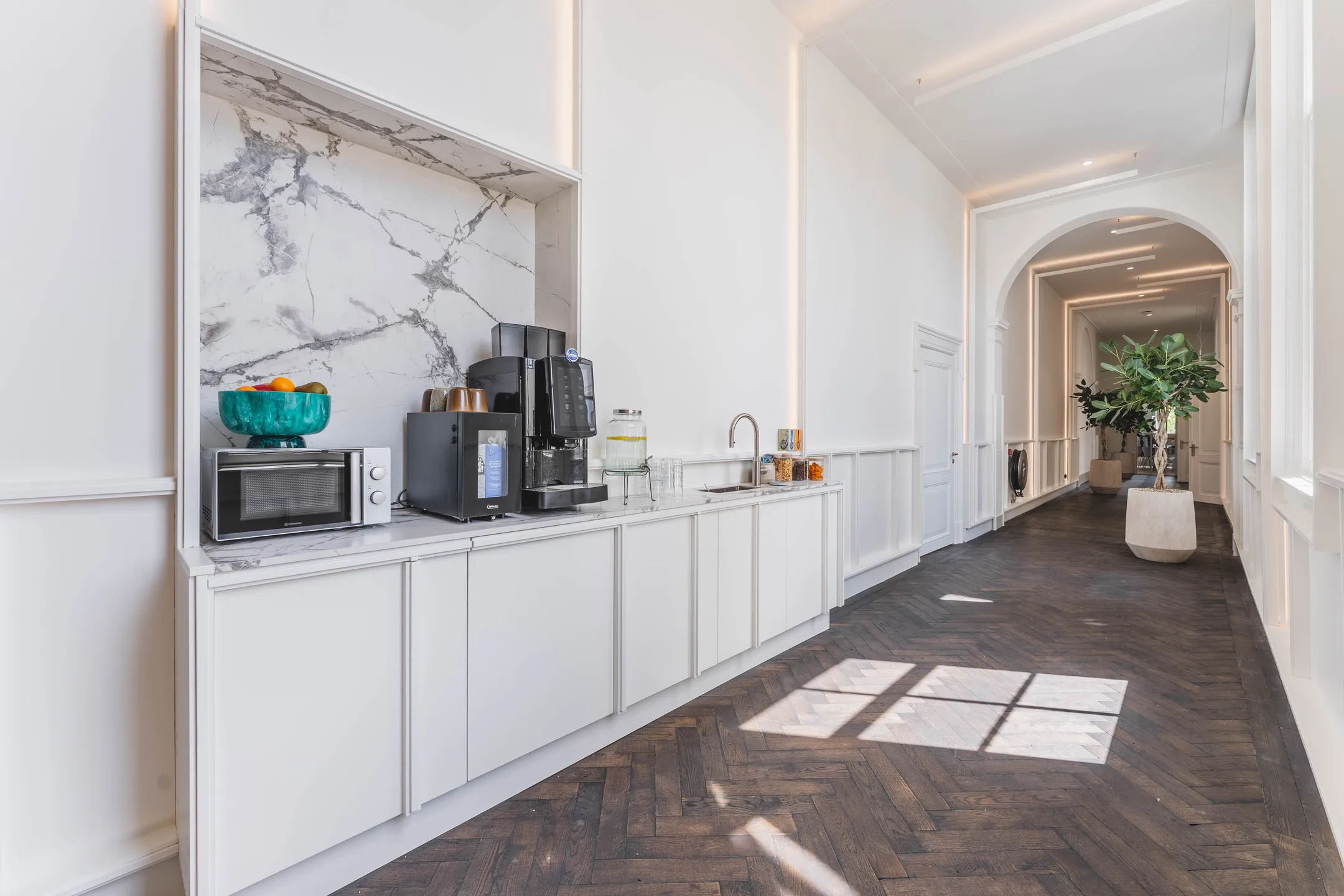 Een lichte keukenhoek met een koffiezetapparaat en magnetron in een gang met houten vloer en planten.