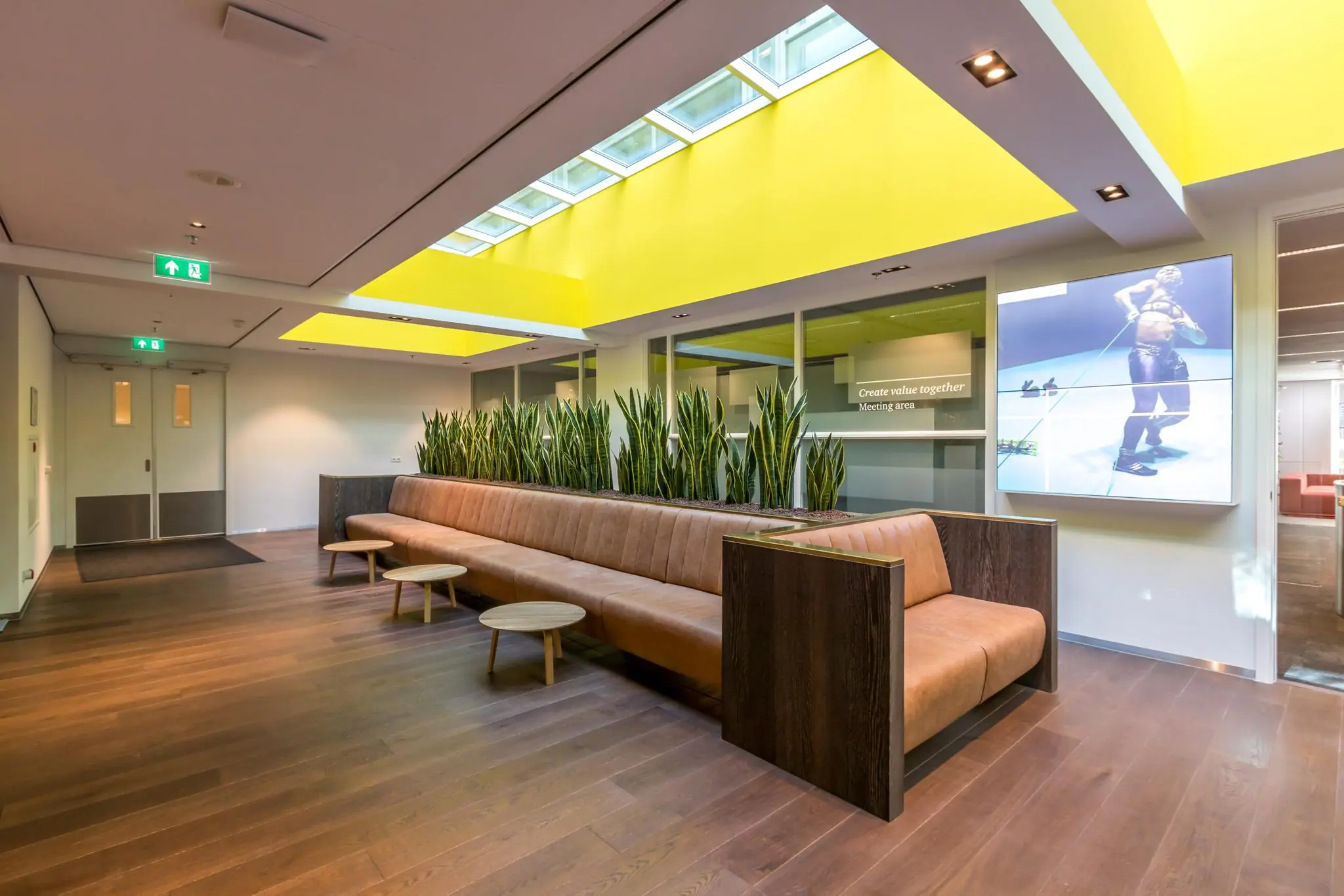 Moderne wachtruimte in het Newtonlaan-kantoorgebouw met lange leren bank, plantenwand en opvallend geel plafond met dakramen.