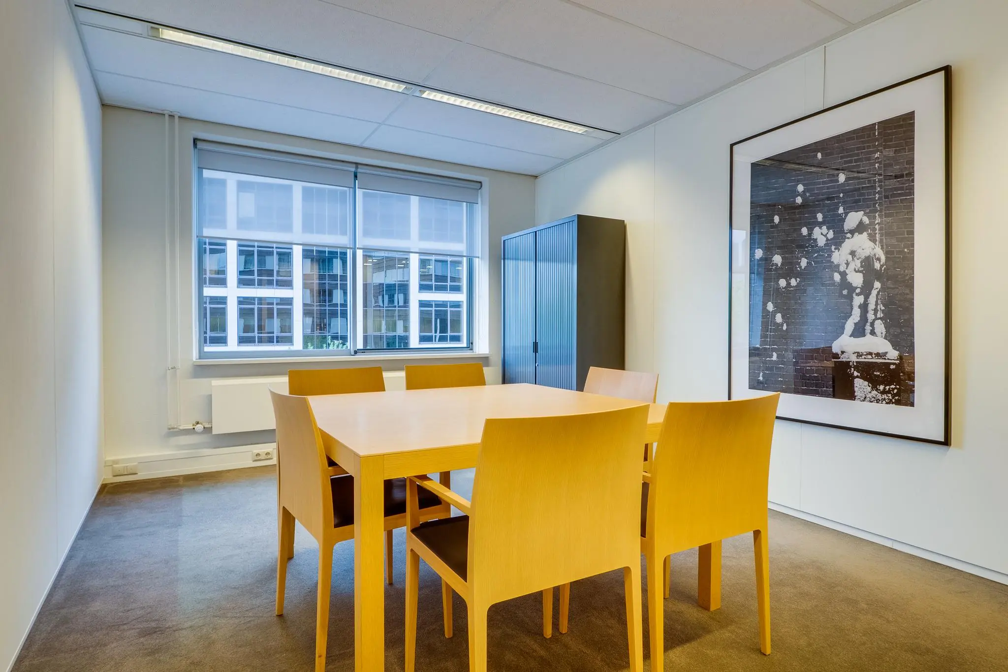 Vergaderruimte met houten tafel en zes stoelen, grote kunstfoto aan de muur en raam met uitzicht op moderne kantoorgebouwen aan de Euclideslaan.