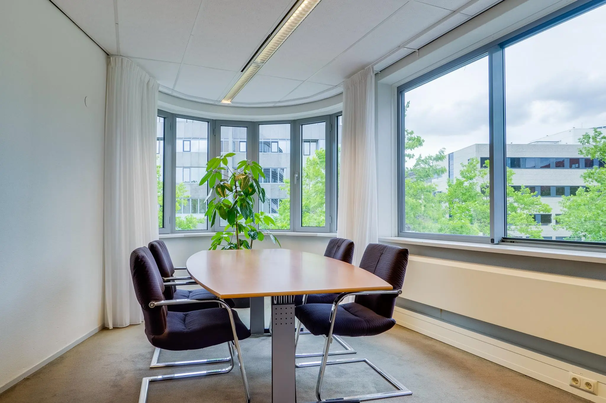 Kleine vergaderruimte aan de Euclideslaan met ovale tafel, vier stoelen en grote ramen met uitzicht op kantoorgebouwen.