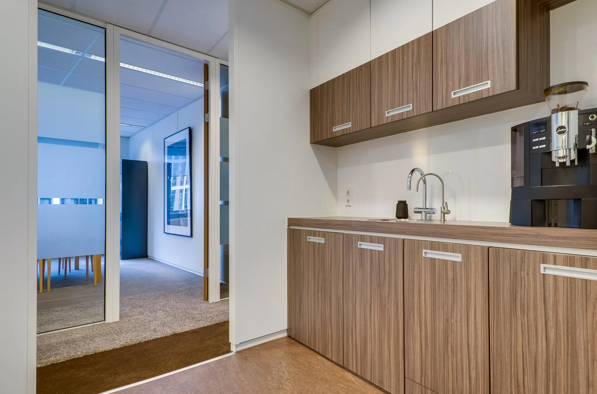 Moderne kantoorpantry met houten keukenkastjes, een koffieautomaat en uitzicht op een vergaderruimte aan de Euclideslaan.