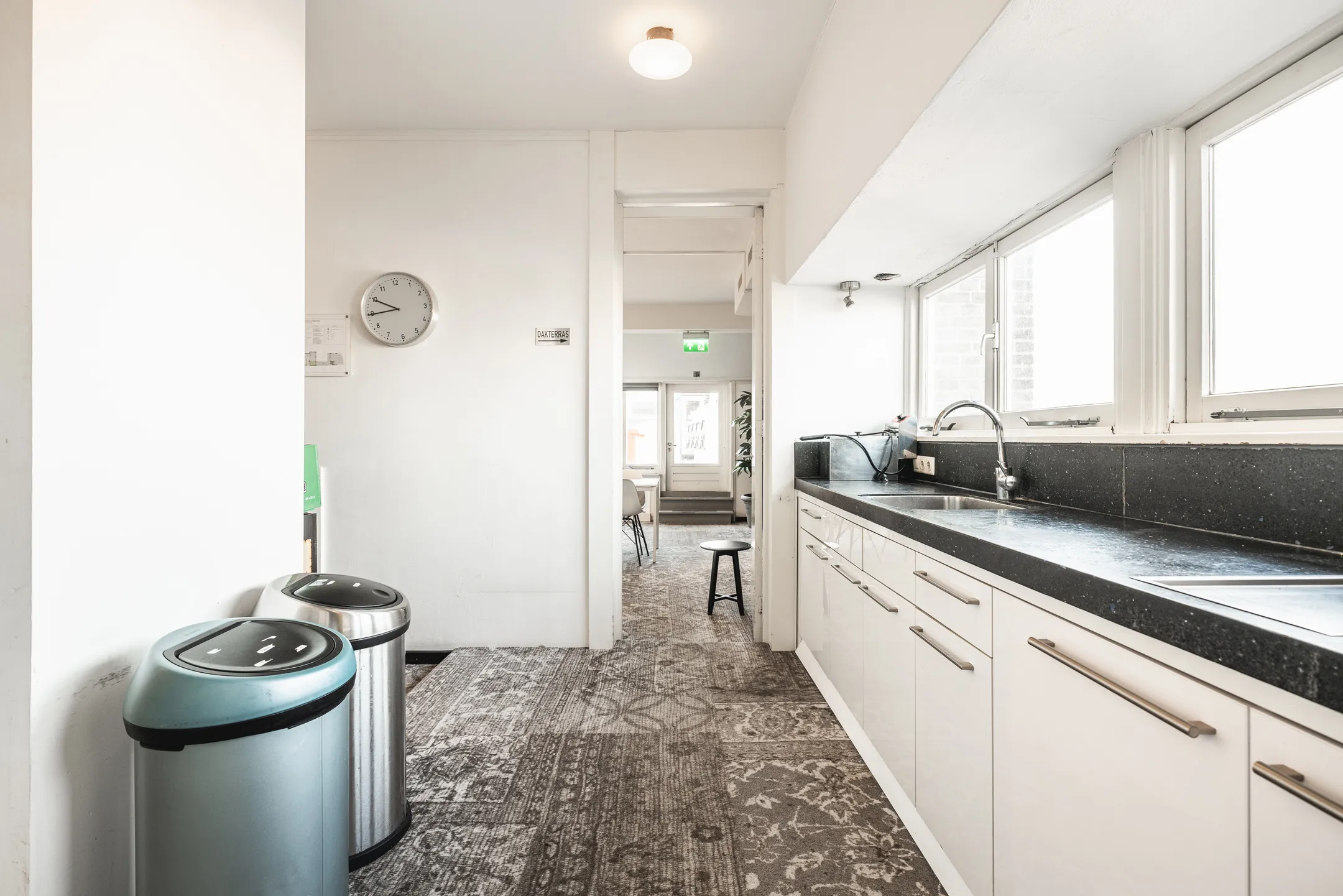 Een lichte keuken met een donker aanrechtblad, klok aan de muur en dubbele prullenbakken.