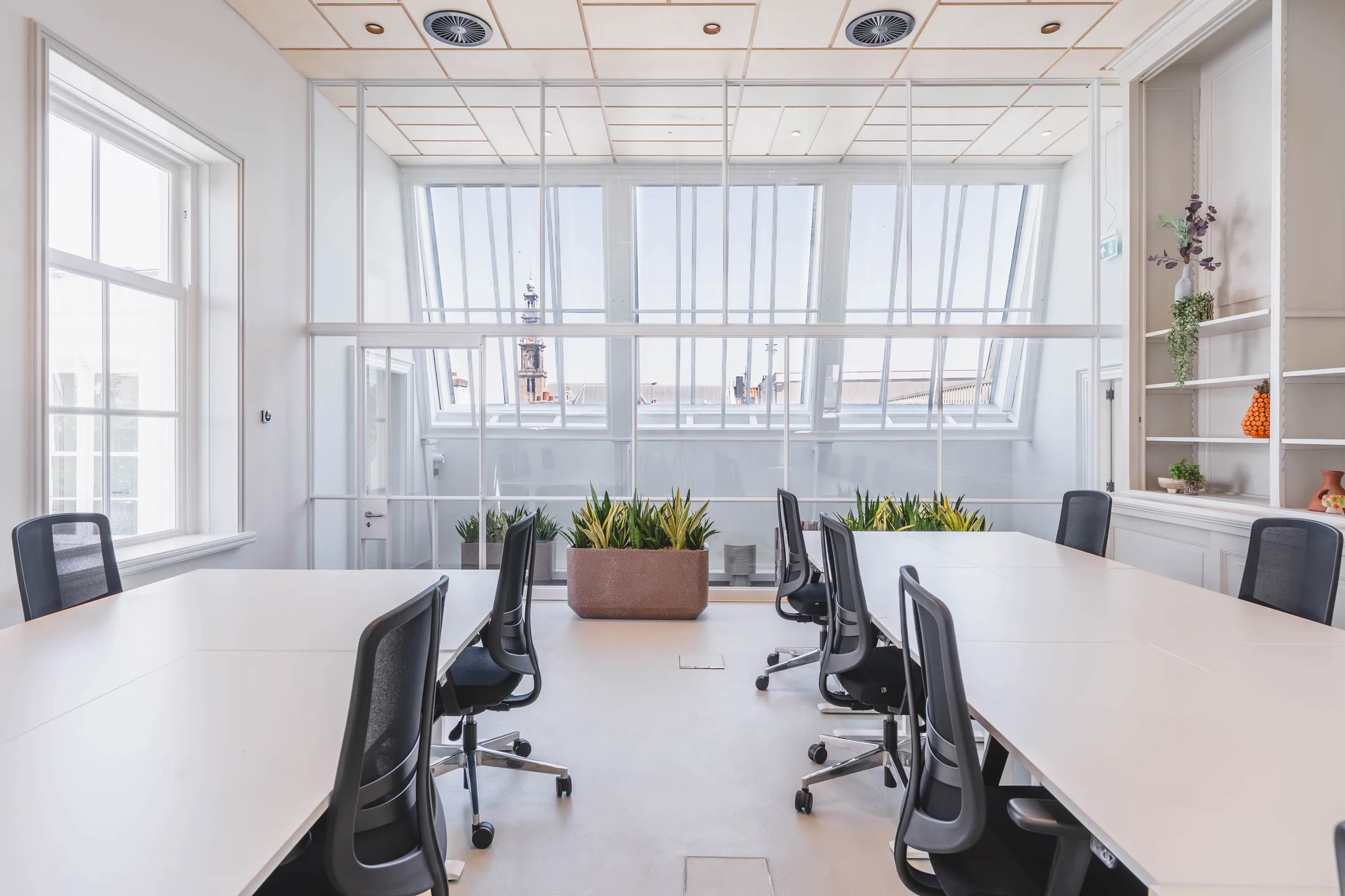 Heldere vergaderruimte met witte tafels, moderne stoelen en grote ramen die veel natuurlijk licht binnenlaten.