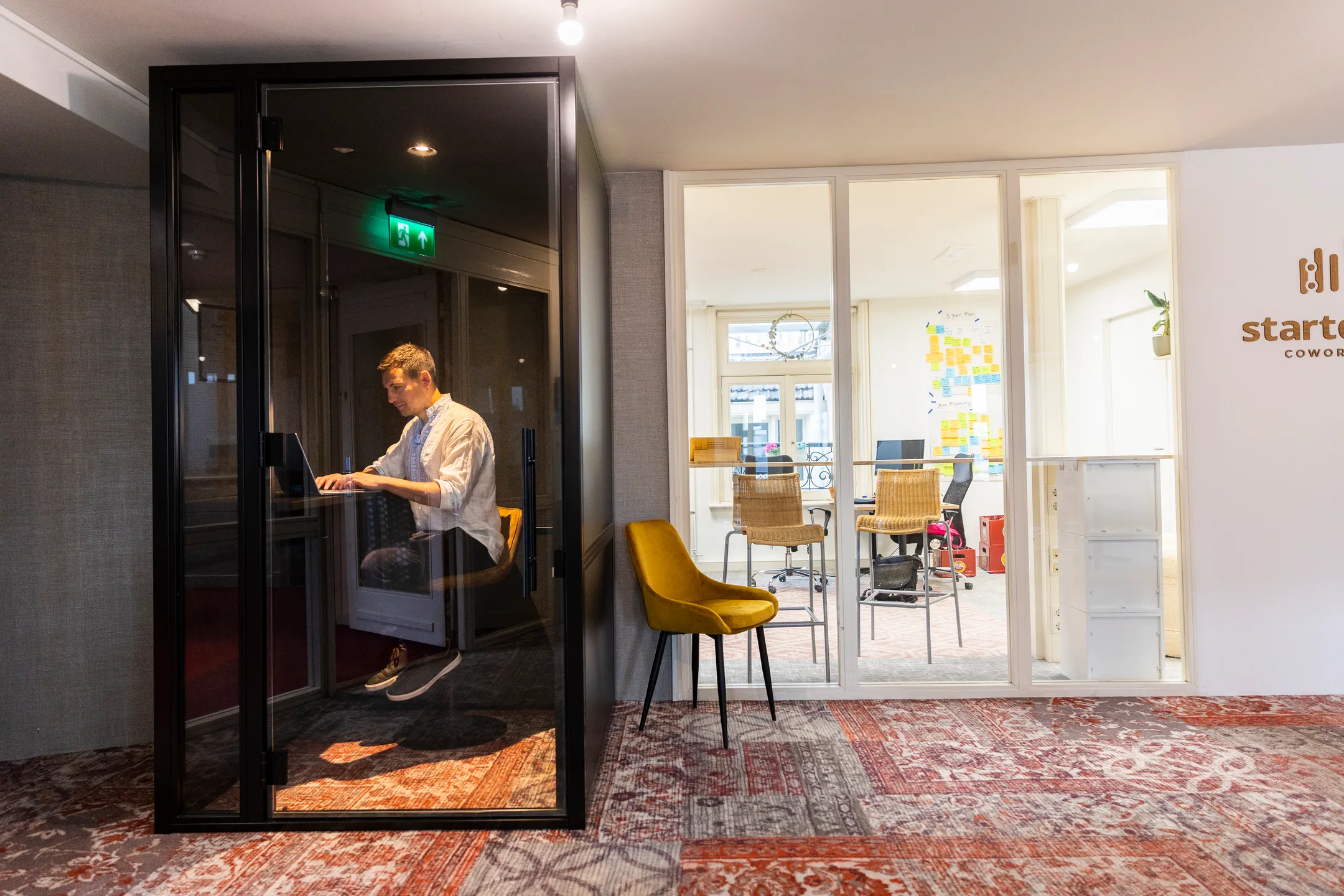 Een man werkt op zijn laptop in een glazen cabine in een modern kantoor.