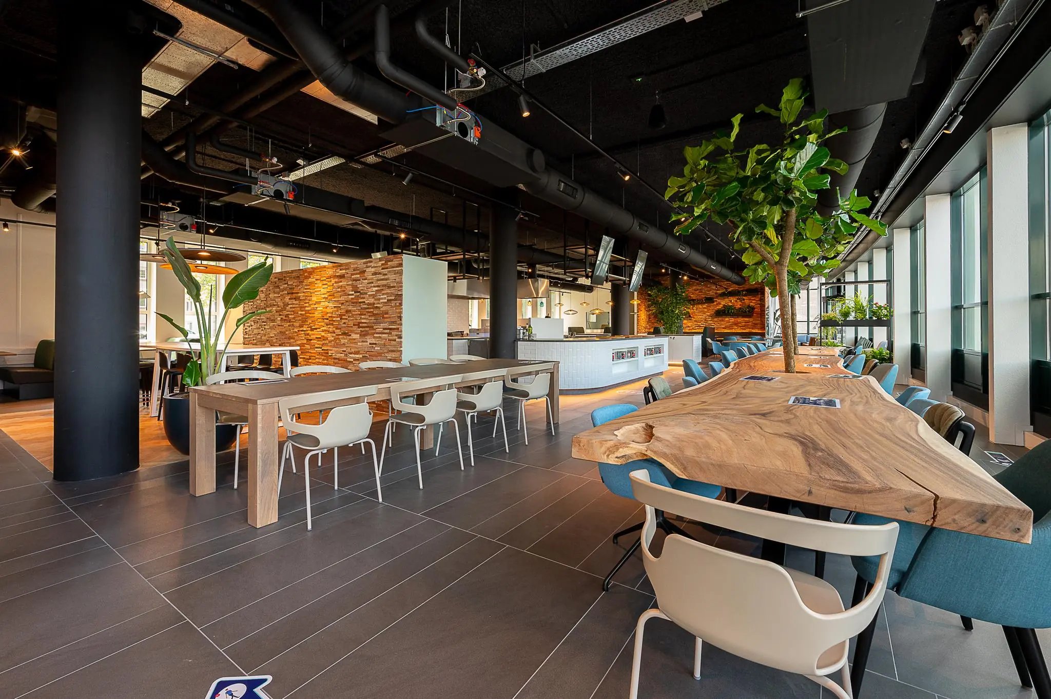 Moderne kantoorkantine aan de Watermanweg met houten tafels, groene planten en een open keuken.