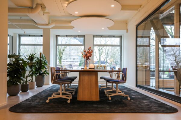Moderne vergaderruimte met houten tafel, acht stoelen, grote ramen en planten in een lichte kantooromgeving.