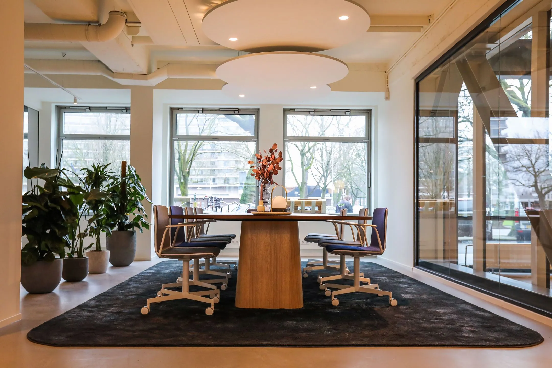 Moderne vergaderruimte met houten tafel, acht stoelen, grote ramen en planten in een lichte kantooromgeving.