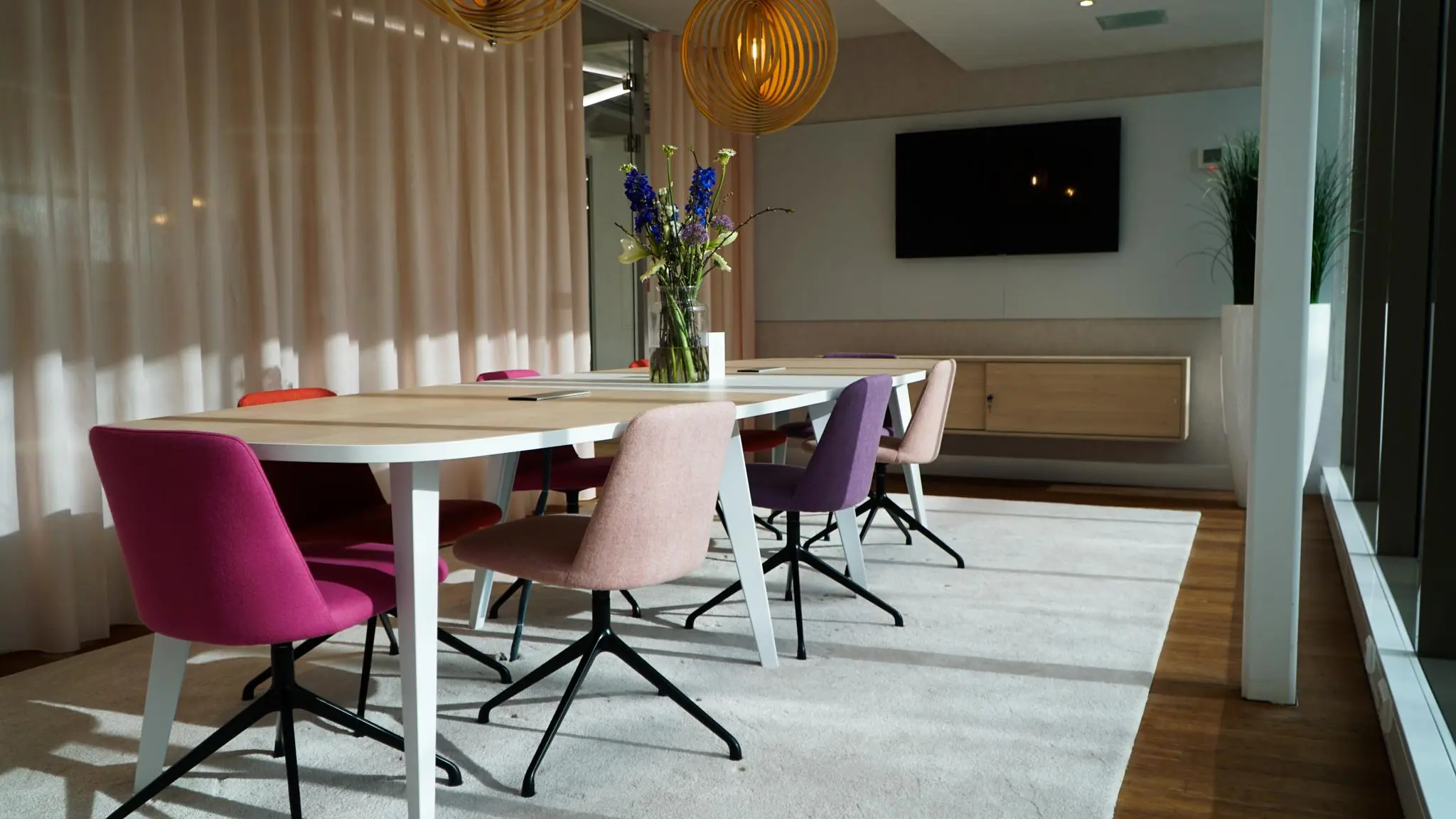 Moderne vergaderruimte met kleurrijke stoelen, een houten tafel, een vaas bloemen en een flatscreen aan de muur.